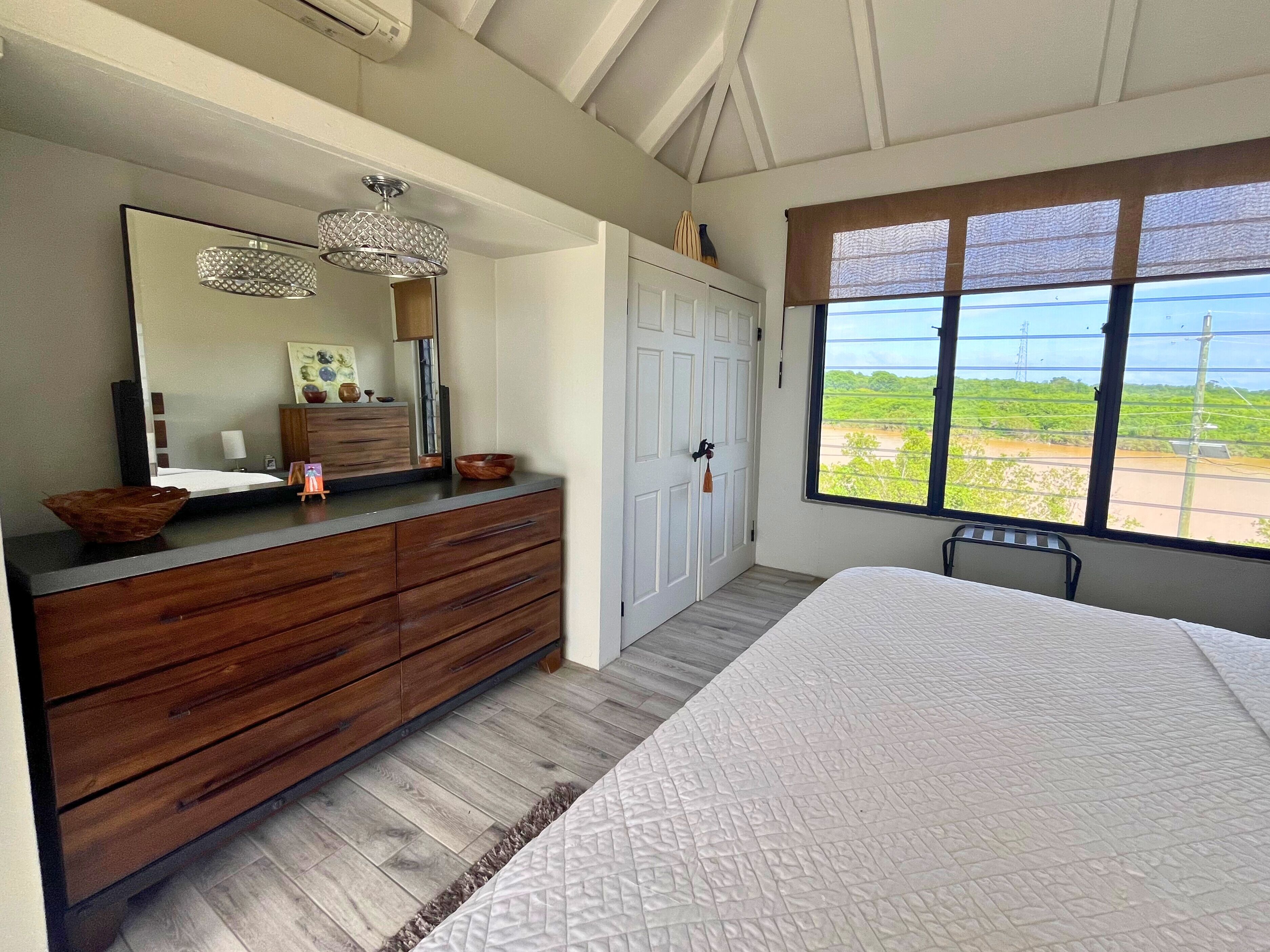 Un dormitorio con una cama grande, un aparador de madera y un espejo amplio. Se aprecia una ventana con vistas a zonas verdes y una silla.