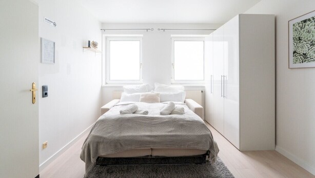 Een minimalistische slaapkamer met een bed, twee ramen, een kledingkast en een lijst met een foto aan de muur.