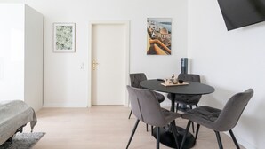 Een moderne, minimalistische kamer met een ronde eettafel, vier gestoffeerde grijze stoelen, een flatscreen televisie aan de muur en twee ingelijste kunstwerken.
