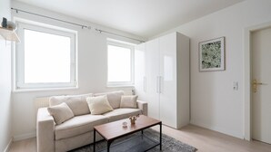 Een moderne woonkamer met een beige bank, een houten salontafel en een grijze vloerkleed. Er zijn twee ramen die zorgen voor natuurlijk licht, een witte deur en een ingelijste botanische print aan de muur.