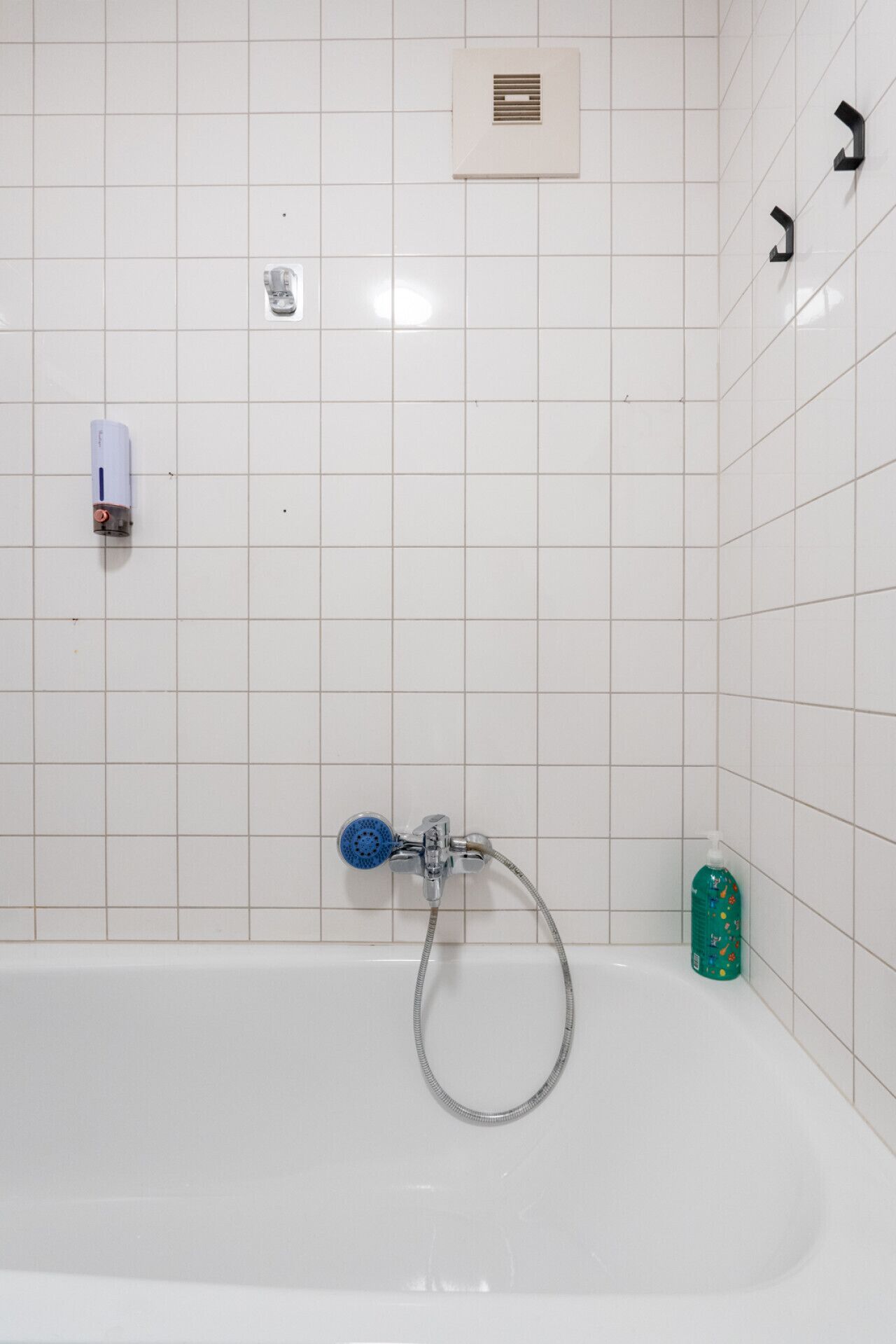 Un baño con una bañera blanca, un cabezal de ducha manual, un dispensador de jabón y una rejilla de ventilación en la pared.