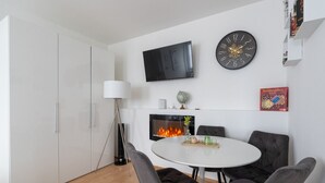 A modern living room with a round dining table, grey upholstered chairs, a flat-screen TV, a decorative clock, a fireplace, and a bookshelf. - Spacious Comfort | 55m² | 6 Guests | 24/7 Check-in (Steyr)