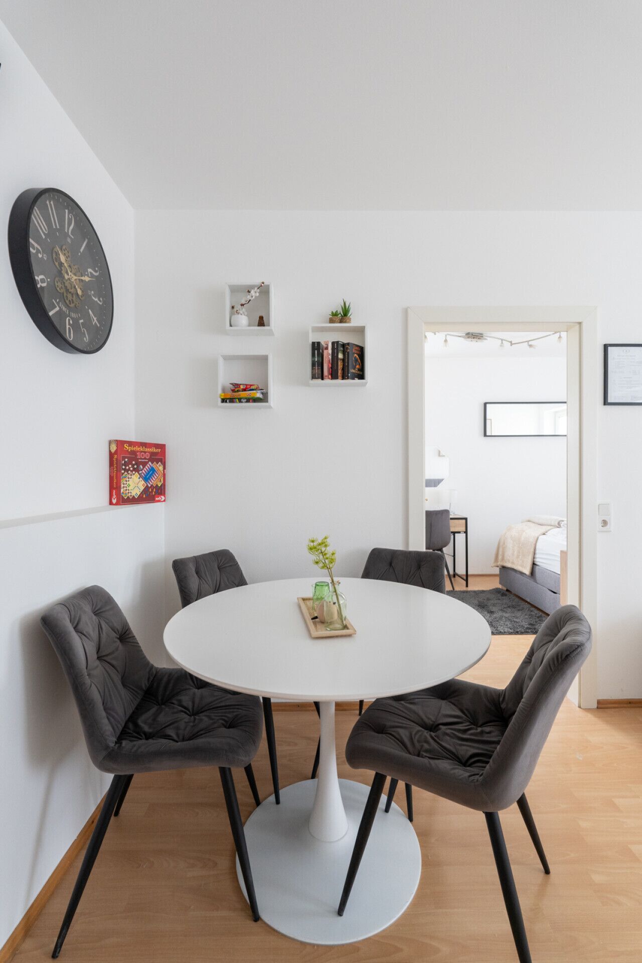 Un comedor moderno con una mesa redonda blanca, cuatro sillas grises tapizadas y un reloj negro en la pared.