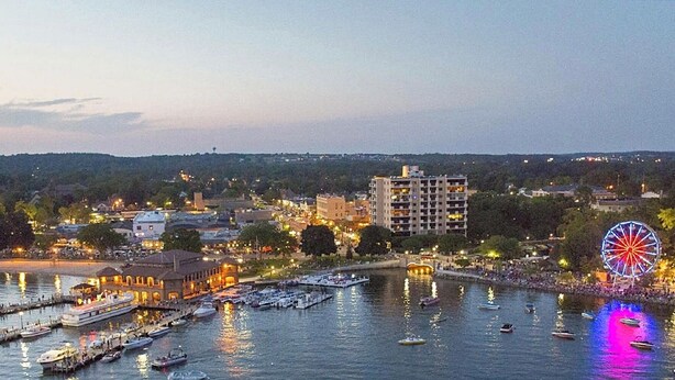 Un paisaje urbano frente al agua, con una noria, barcos amarrados al muelle y edificios al fondo.