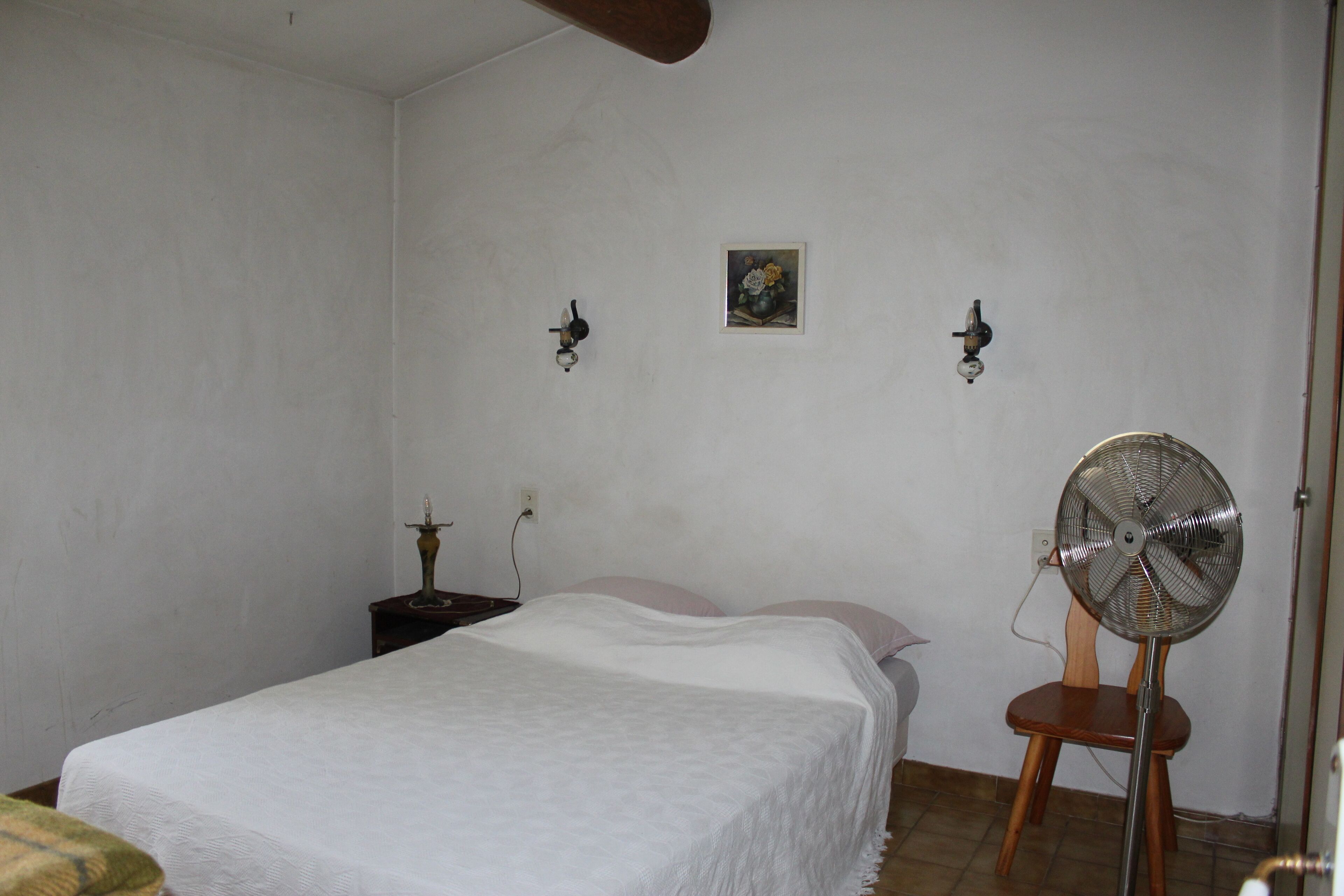 A bedroom with a bed, a wooden chair, a standing fan, and a small table with a lamp.