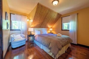 A bedroom with a wooden sloped ceiling, a bed with a beige comforter, two bedside lamps, a nightstand, and a chair.