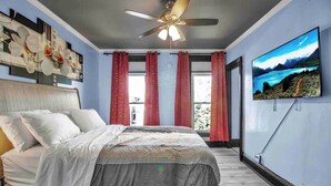 A bedroom with a bed, a ceiling fan, a large window with red curtains, and a wall-mounted TV displaying a scenic landscape. - Spacious 6-bedroom house with WiFi, AC  and free Parking in brilliant Buffalo (Buffalo)