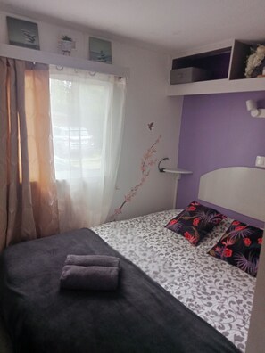 A small, neatly arranged bedroom with a bed, a window with sheer curtains, a purple wall, and a shelf above the bed.
