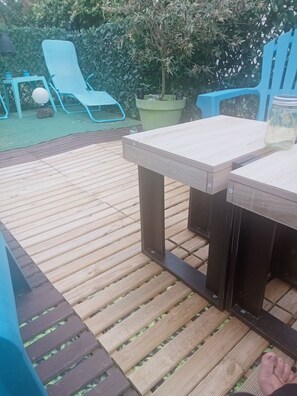 A wooden deck with a table and chairs, surrounded by greenery and a blue fence.
