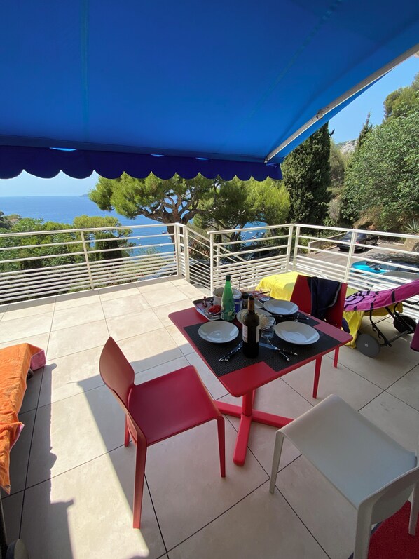 Eine Terrasse im Freien mit einem für zwei Personen gedeckten Tisch, umgeben von roten Stühlen, unter einer blauen Markise.