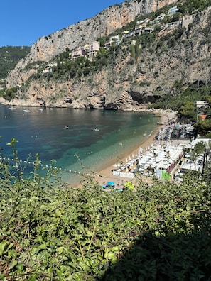Eine Küstenlandschaft mit einer felsigen Klippe, einem Strand mit Sonnenschirmen und Booten auf dem Wasser.