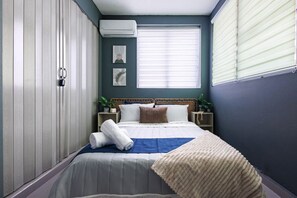 A bedroom with a bed, bedside tables, a wall-mounted air conditioner, and a window with blinds.