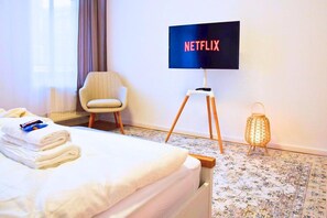 Habitación de hotel con cama, una silla, televisión sobre un soporte, lámpara y radiador.