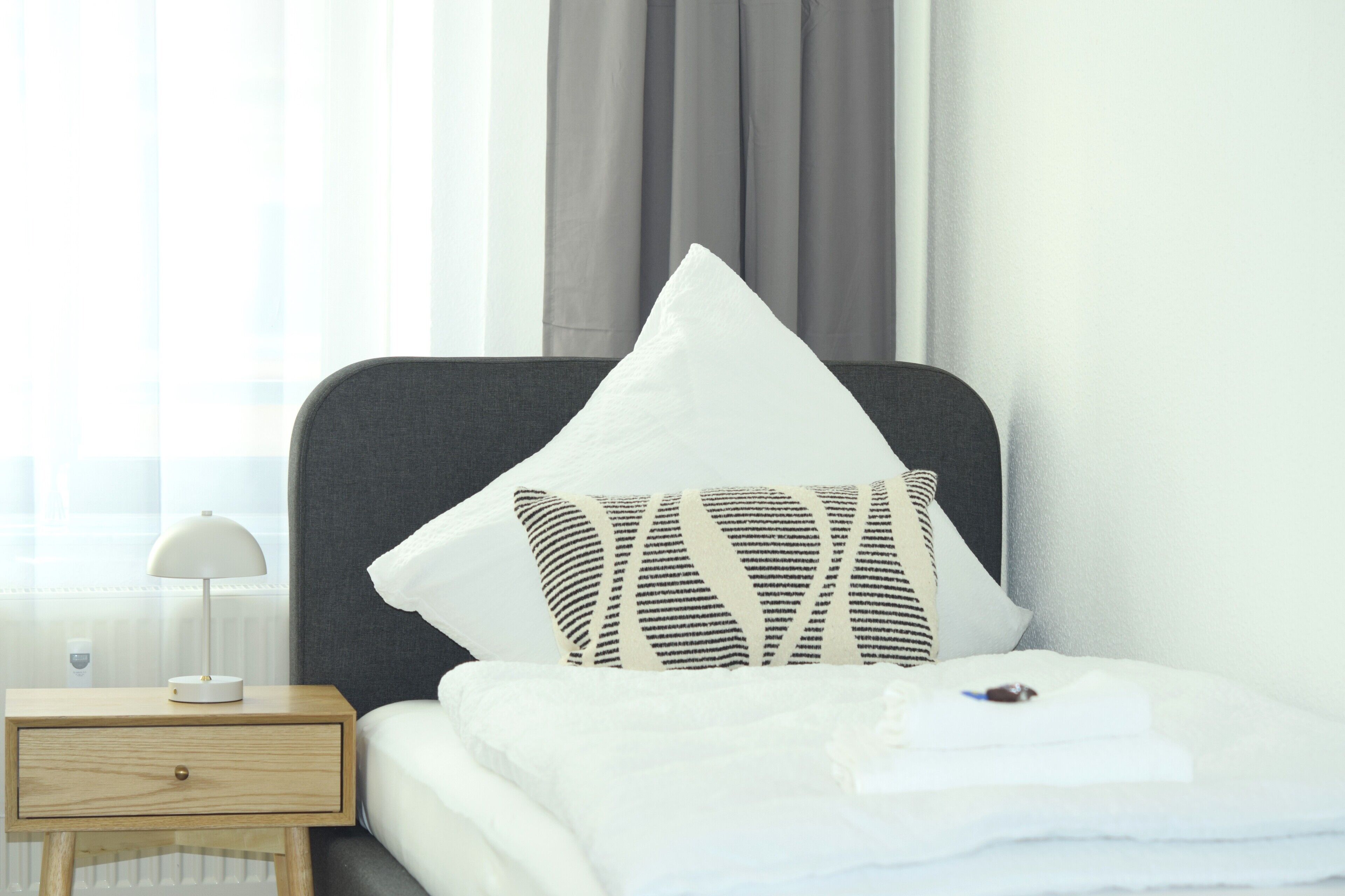 Ein ordentlich bezogenes Bett mit weißer Bettwäsche und einem Kopfkissen mit Streifenmuster. Ein kleiner Nachttisch mit einer Lampe steht daneben.