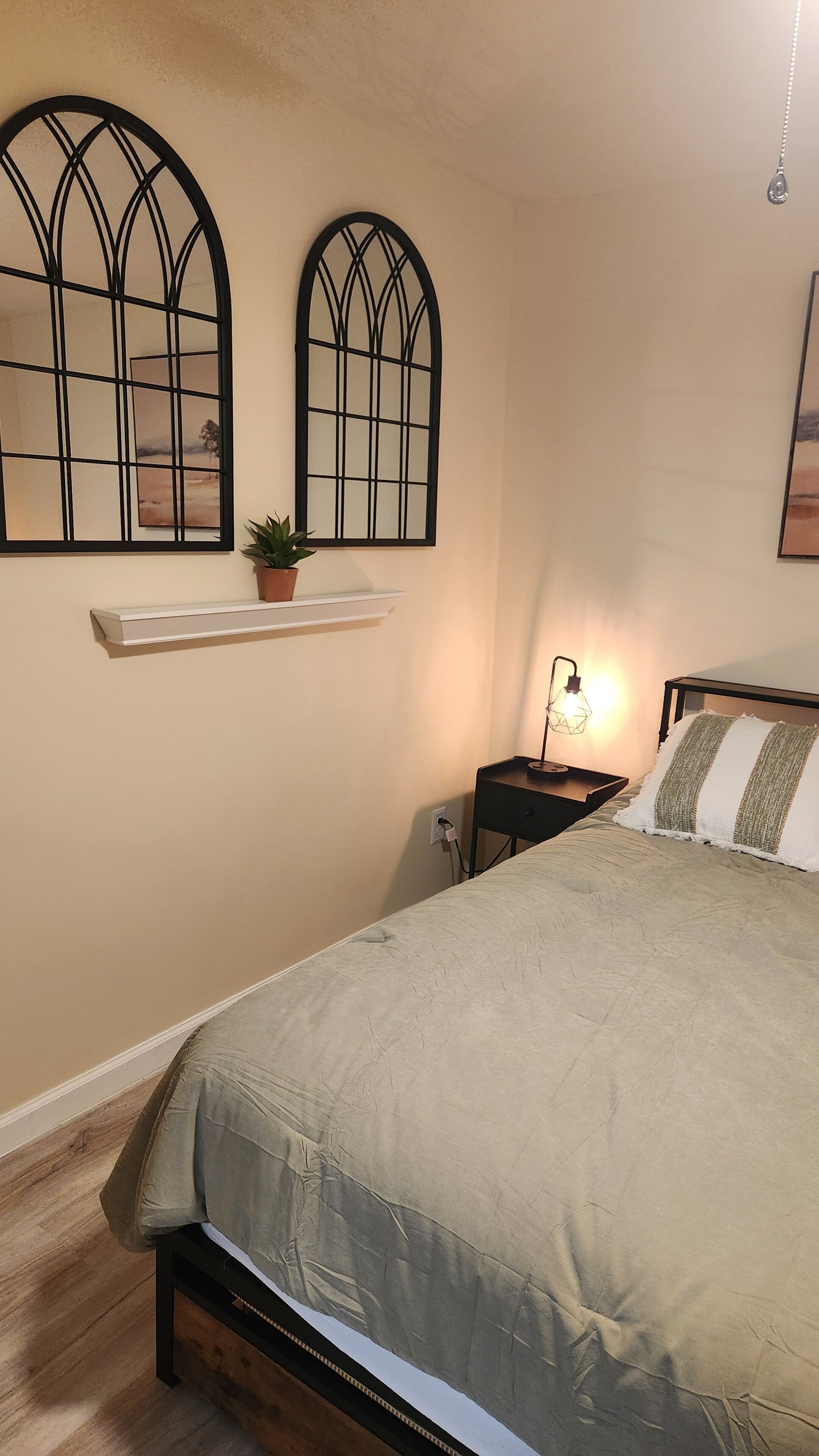 A bedroom with a bed, a nightstand, a wall-mounted shelf with a plant, and two arched windows.