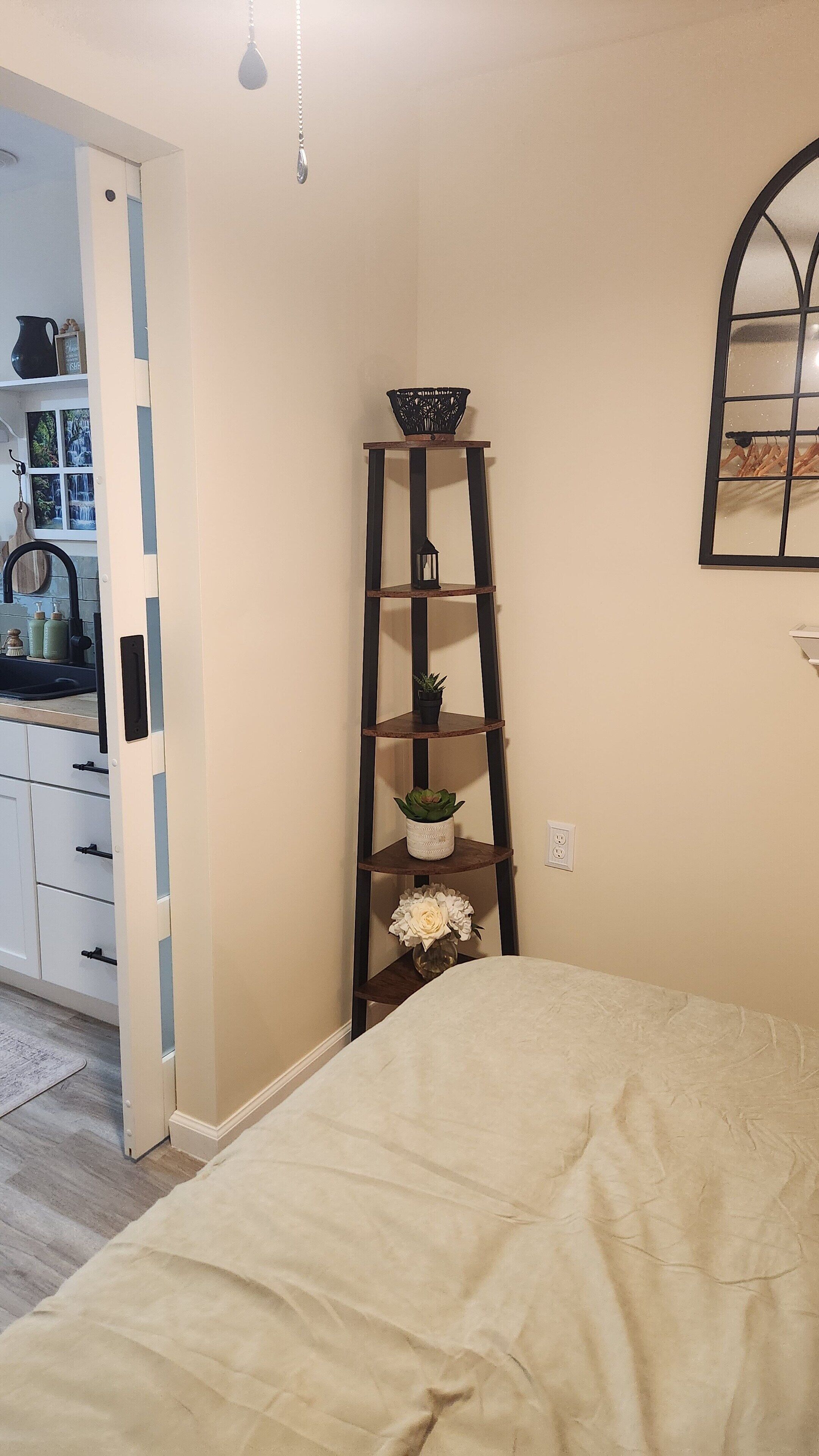 A bedroom with a bed, a wooden ladder shelf with plants, a window with a view, and a door leading to a bathroom.
