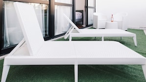 Ett modernt utomhusområde med bekväma vita loungestolar och ett litet bord, placerat framför stora fönster och en grön konstgräsmatta.