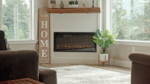 Ein Wohnzimmer mit Kamin, einem darüber hängenden Fernseher und einem großen Fenster mit Blick auf Bäume.