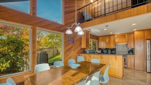 Une cuisine moderne avec des meubles en bois, un coin repas avec une table en bois et des chaises bleu clair, et une vue sur un jardin grâce à de grandes fenêtres.