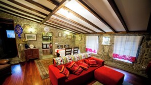 A cozy room with a red sofa, wooden beams, stone walls, a dining area with chairs, and a TV.