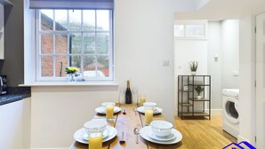 A modern kitchen with a dining table set for breakfast, a window with a view of a brick building, and a washing machine in the adjacent room. - 2br-sleeps4-citycentre-freeparking (Derby)