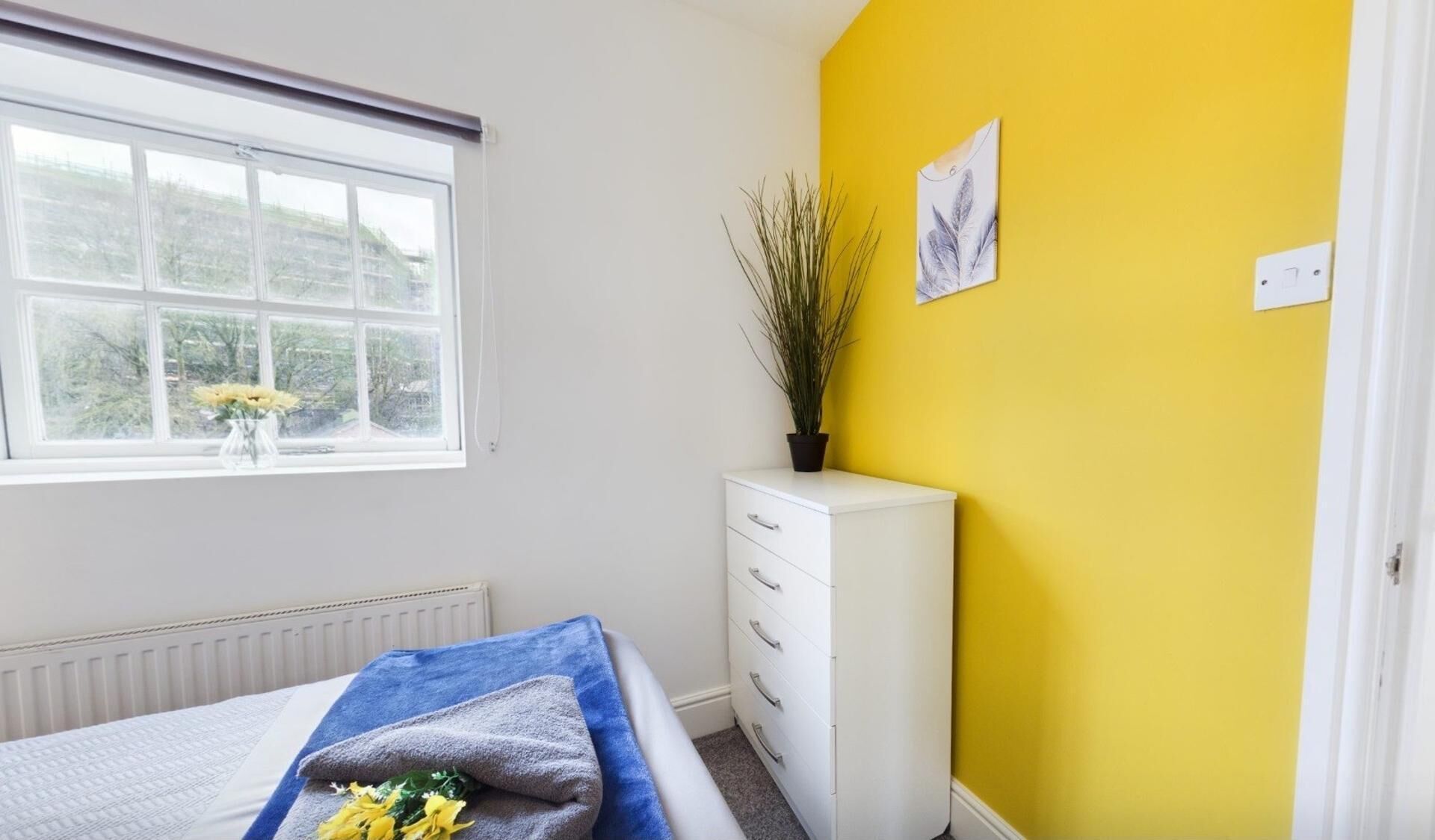 A bedroom with a bed, a window, a radiator, a white dresser, and a yellow wall.