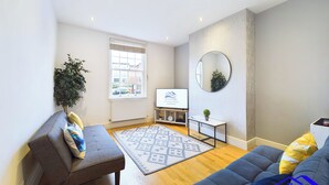A modern living room with a grey sofa, a blue sofa, a patterned rug, a small table with a plant, and a TV displaying a company logo. - 2br-sleeps4-citycentre-freeparking (Derby)