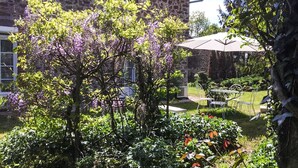 A stone building with a garden in front, including a tree with purple flowers and a patio area with chairs and an umbrella.