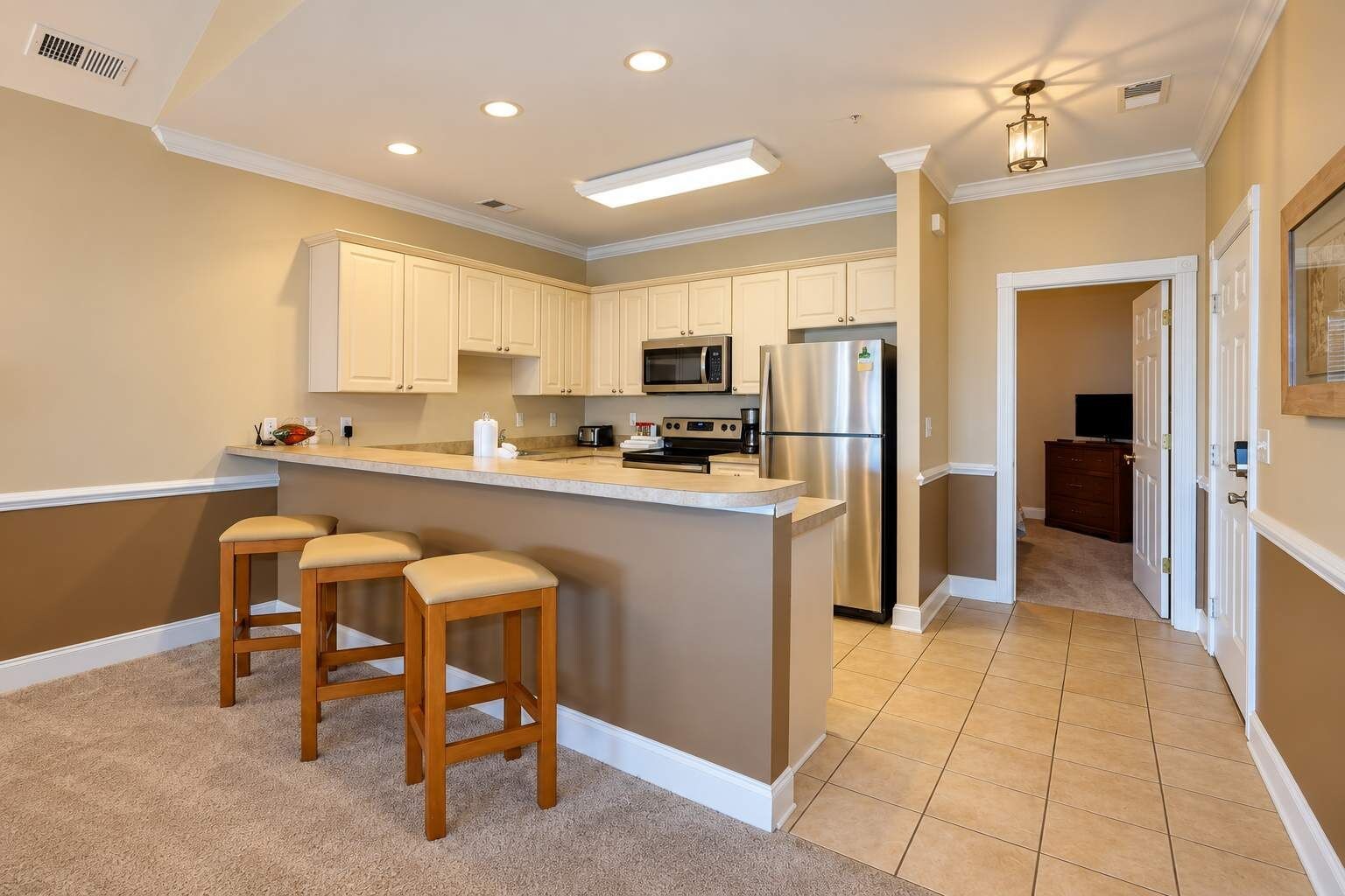 A kitchen with white cabinets, a stainless steel refrigerator, a microwave, and a bar stool area.