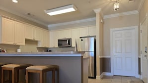 A kitchen with white cabinets, a stainless steel refrigerator, a microwave, and a bar stool area. - Myrtlewood Condo-Covered Porch, Pool & Hot Tub (Myrtle Beach)