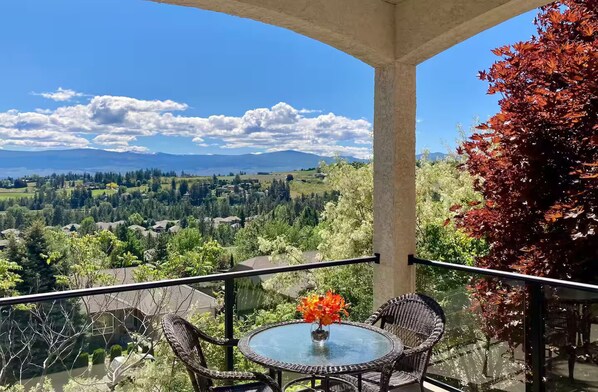 Un balcon doté de meubles en osier et d’une table en verre, offrant une vue panoramique sur un paysage pittoresque, avec des maisons et des montagnes.