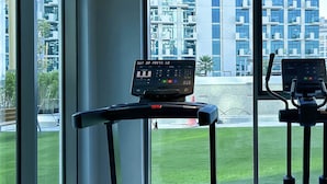 Une salle de sport équipée d’un tapis de course et d’un vélo elliptique, de grandes fenêtres offrant une vue sur un bâtiment moderne et une pelouse verdoyante.