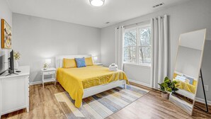 Une chambre à coucher avec un lit, des tables de chevet, une télévision, un miroir et une fenêtre avec des rideaux.