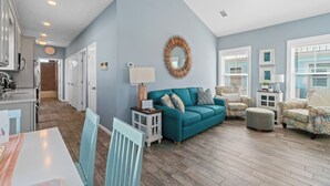A spacious living room with a teal sofa, patterned armchairs, and a ceiling fan. A kitchen is visible in the background with white cabinets and a dining area with a table and chairs.