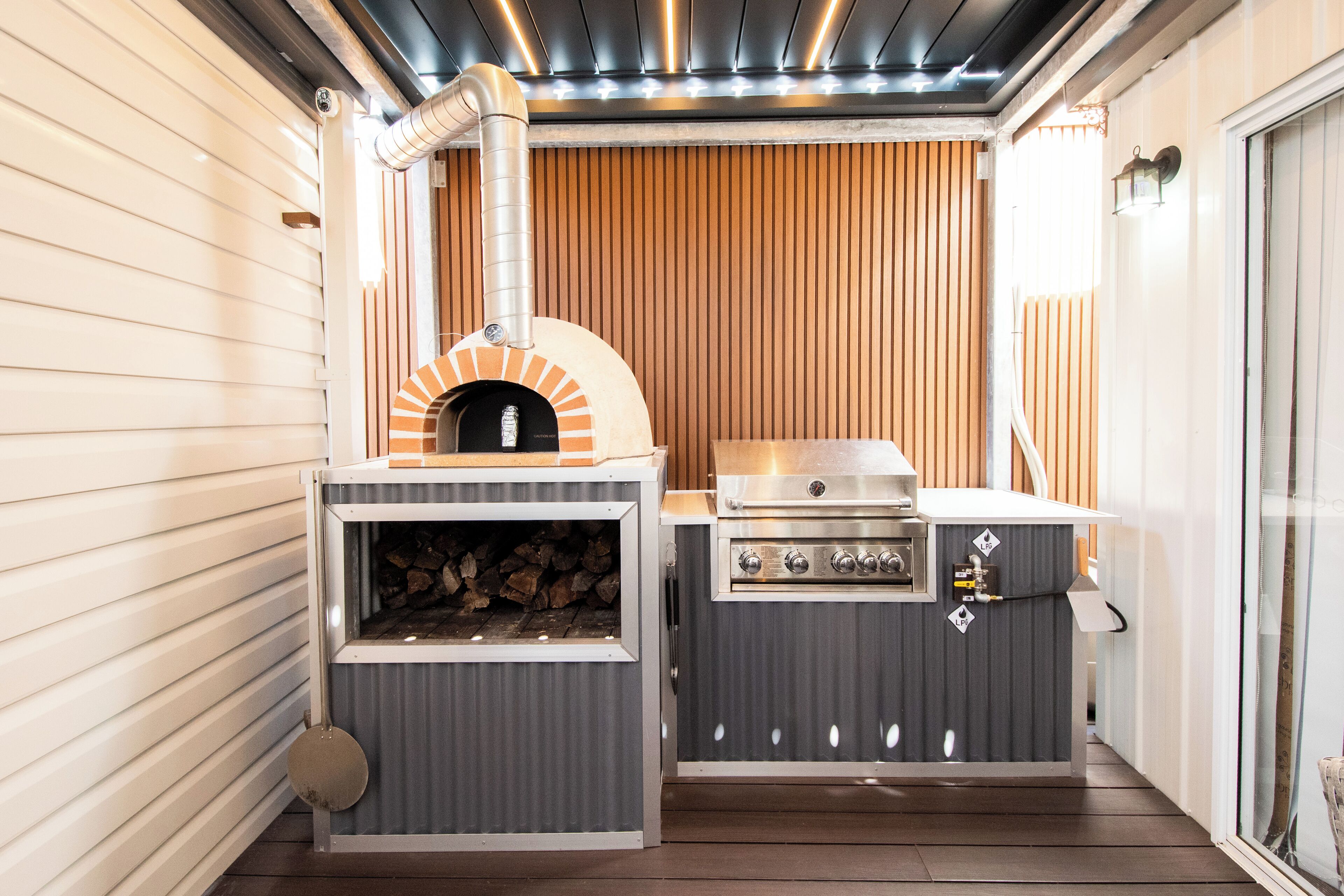 A compact outdoor kitchen with a wood-fired oven, a grill, and a built-in wood storage area.