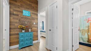 A hallway with wooden flooring, a blue dresser with a vase of flowers, a wall-mounted sign, and a door leading to another room. - Coastal Getaway w/ Community Pool + Fishing Nearby (Rockport)