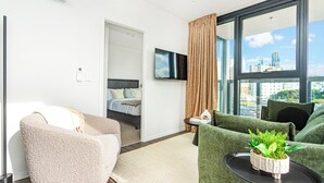 Une chambre d’hôtel moderne avec un canapé, une petite table agrémentée d’une plante, un lit avec une tête de lit, un téléviseur fixé au mur et une grande fenêtre donnant sur la ville.