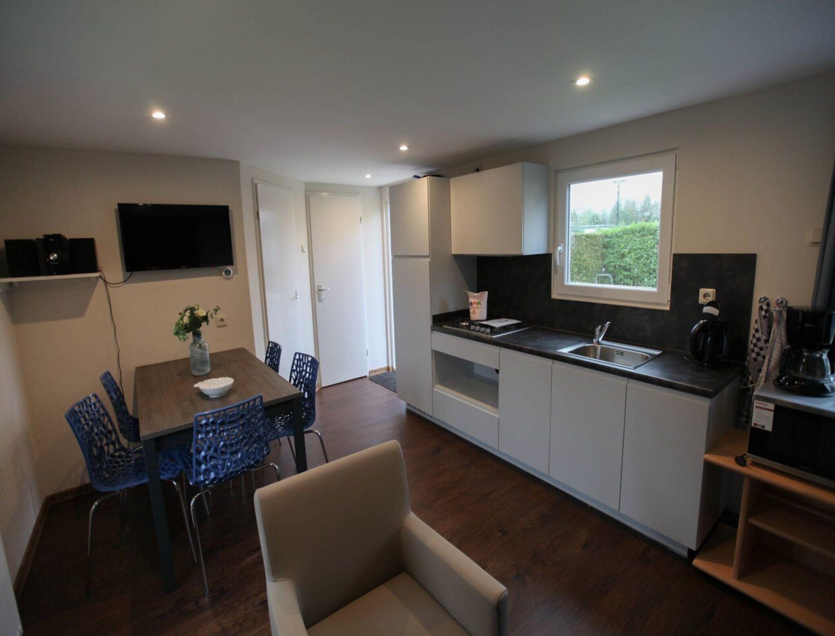 A compact kitchen with a dining area, a TV mounted on the wall, and a window providing a view of greenery outside.