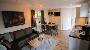 A modern living room with a sofa, coffee table, and dining area. There is a kitchen with a stove and sink. A painting is on the wall, and a window with curtains is visible. - Detached Chalet for 5 People in Terwolde (Terwolde)