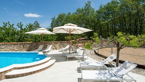 Ein Schwimmbecken mit Liegestühlen und Sonnenschirmen, umgeben von einer Steinmauer und Bäumen.