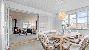 A spacious living area with a round wooden table, white cushioned chairs, a large window with a view, and a built-in wood stove.