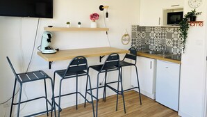 A compact kitchen with a wooden table, four black chairs, a coffee machine, a TV mounted on the wall, and a clock.