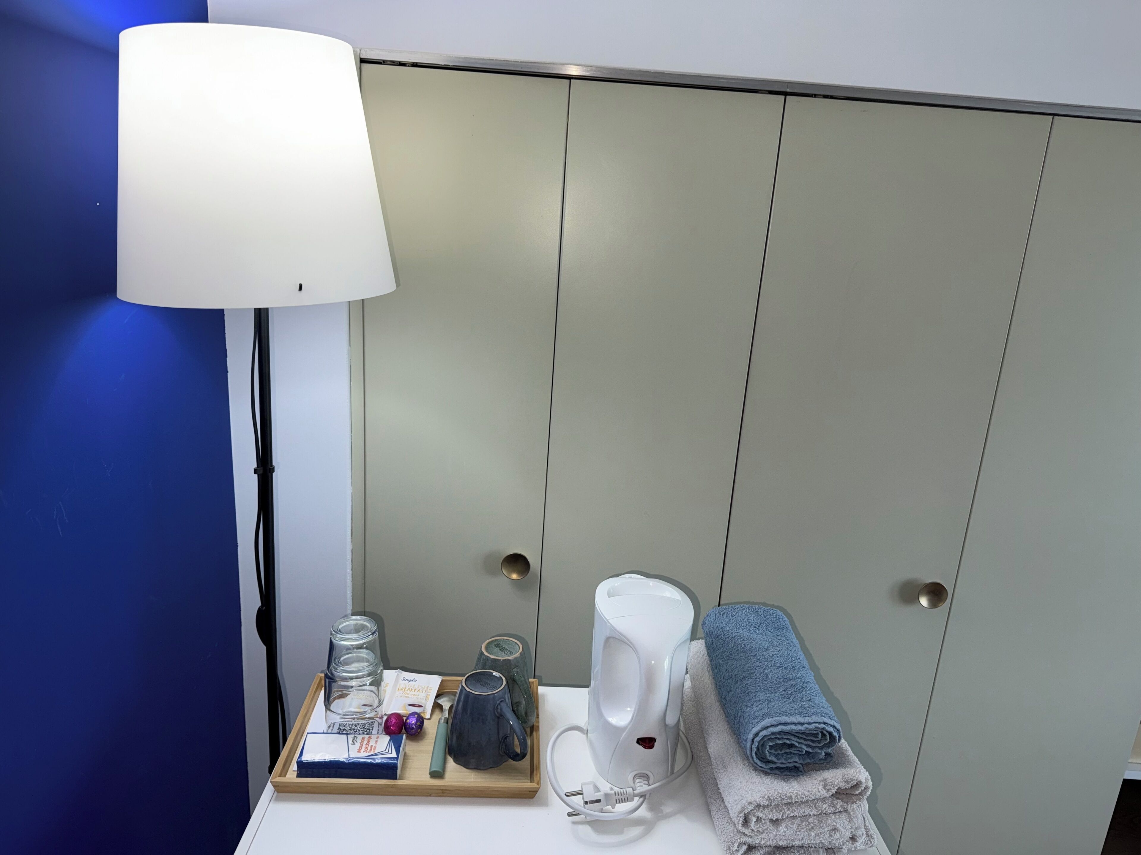 Una habitación de hotel con una mesita de noche que tiene una lámpara, un secador de pelo, una toalla y una bandeja con una taza, vasos y una caja de pañuelos.