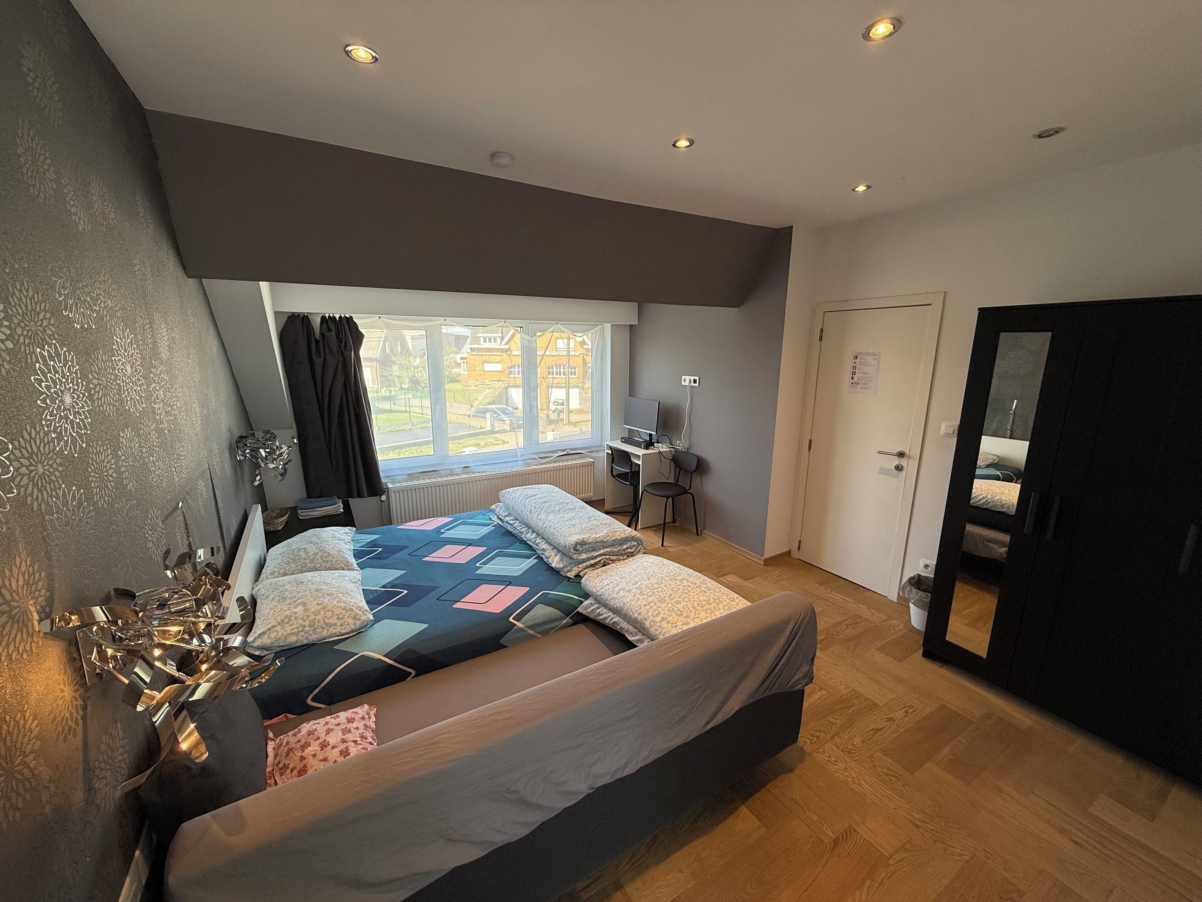 Ein Schlafzimmer mit einem Bett, einem Schreibtisch und einem Computer. Ein Fenster bietet Blick auf Häuser.