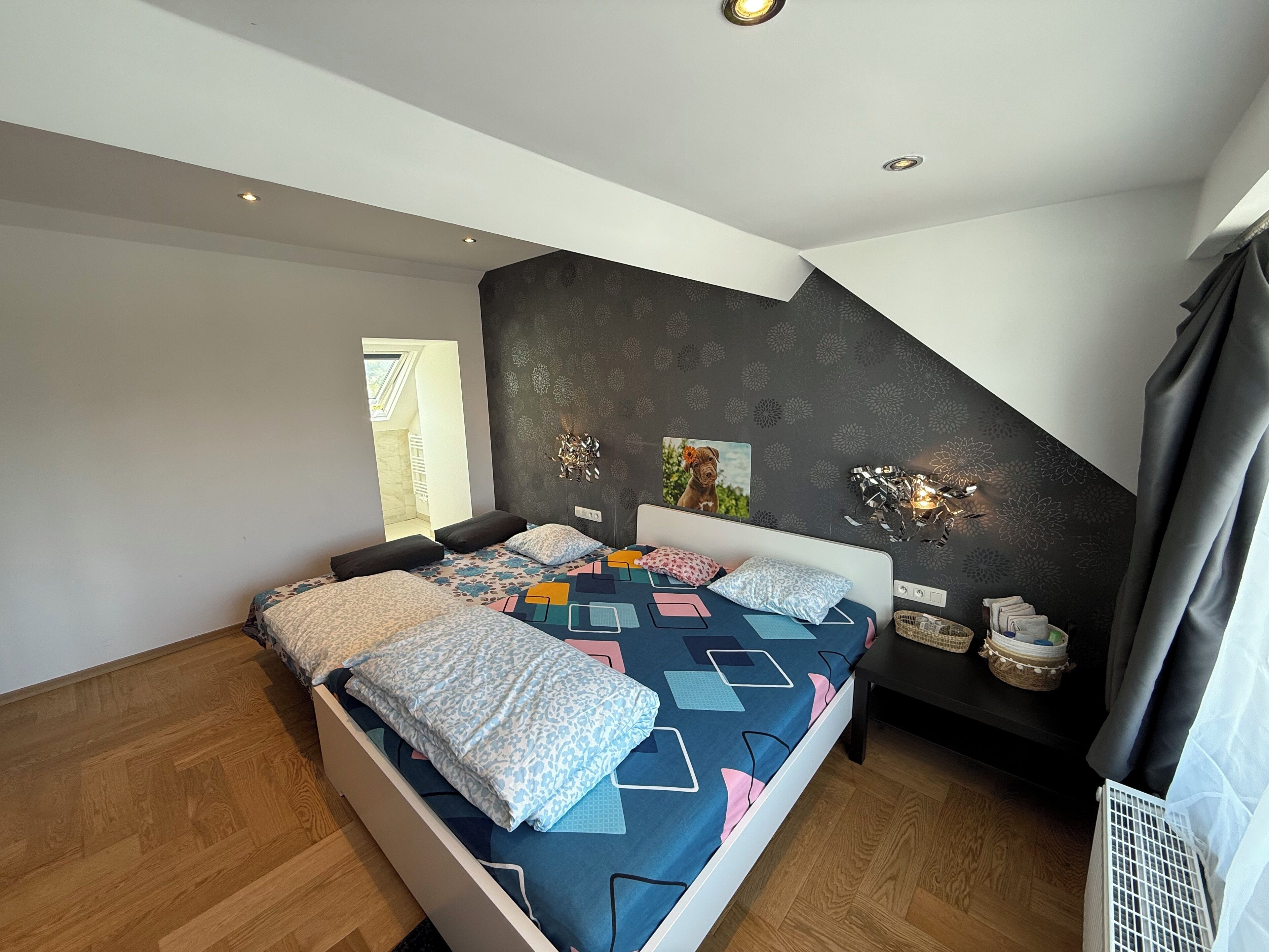 A modern bedroom with a sloped ceiling, a large bed with patterned bedding, a wall-mounted light fixture, and a small side table with decorative items.
