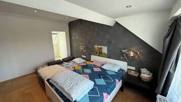 A modern bedroom with a sloped ceiling, a large bed with patterned bedding, a wall-mounted light fixture, and a small side table with decorative items.