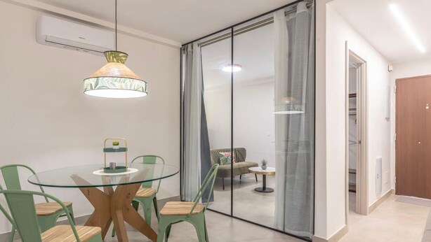 Een moderne eethoek met een glazen tafel en stoelen, een hanglamp en een schuifdeur van glas die leidt naar een woonruimte.