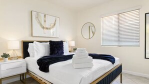 A hotel room with a bed, bedside tables, a window with blinds, and wall art. - Quiet 4 BD Home w/ Pool (North Las Vegas)