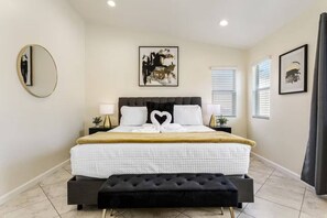 A bedroom with a bed, a black ottoman, a round mirror, and artwork on the walls. - Quiet 4 BD Home w/ Pool (North Las Vegas)