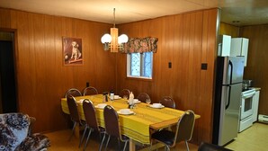 A dining area with a wooden paneled wall, a dining table set with chairs, a chandelier, and a refrigerator in the adjacent kitchen. - Family Reunion Cottage - Can sleep 16+  - Includes Motor Boat, Canoe, AC - Wi-Fi (Britt)